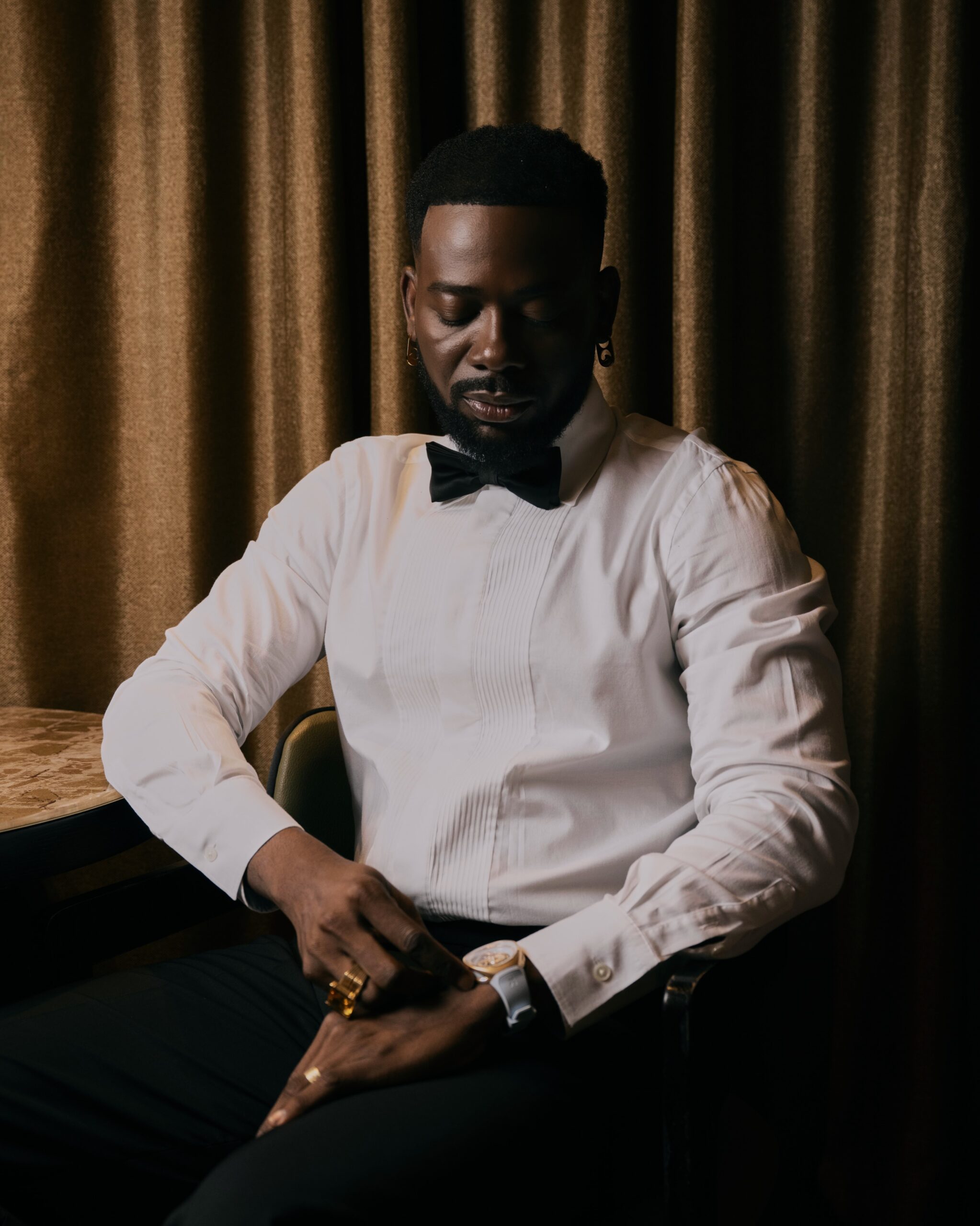 Adekunle Gold wearing a white pleated dress shirt, black satin bow tie, gold ring, and leather strap wristwatch.