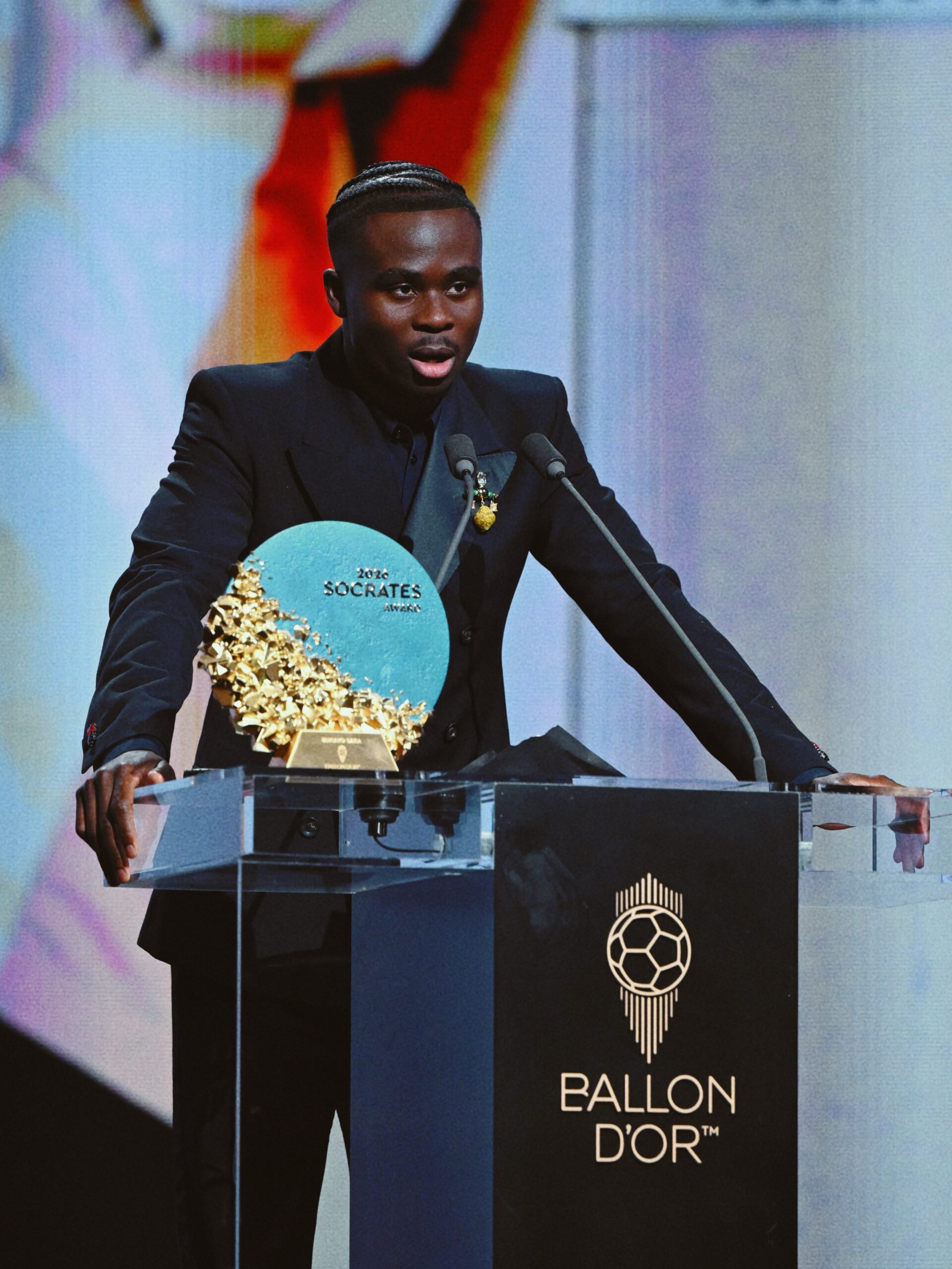 Bukayo Saka standing behind the podium holding the 2026 Socrates Award plaque at the Ballon d’Or ceremony in London.