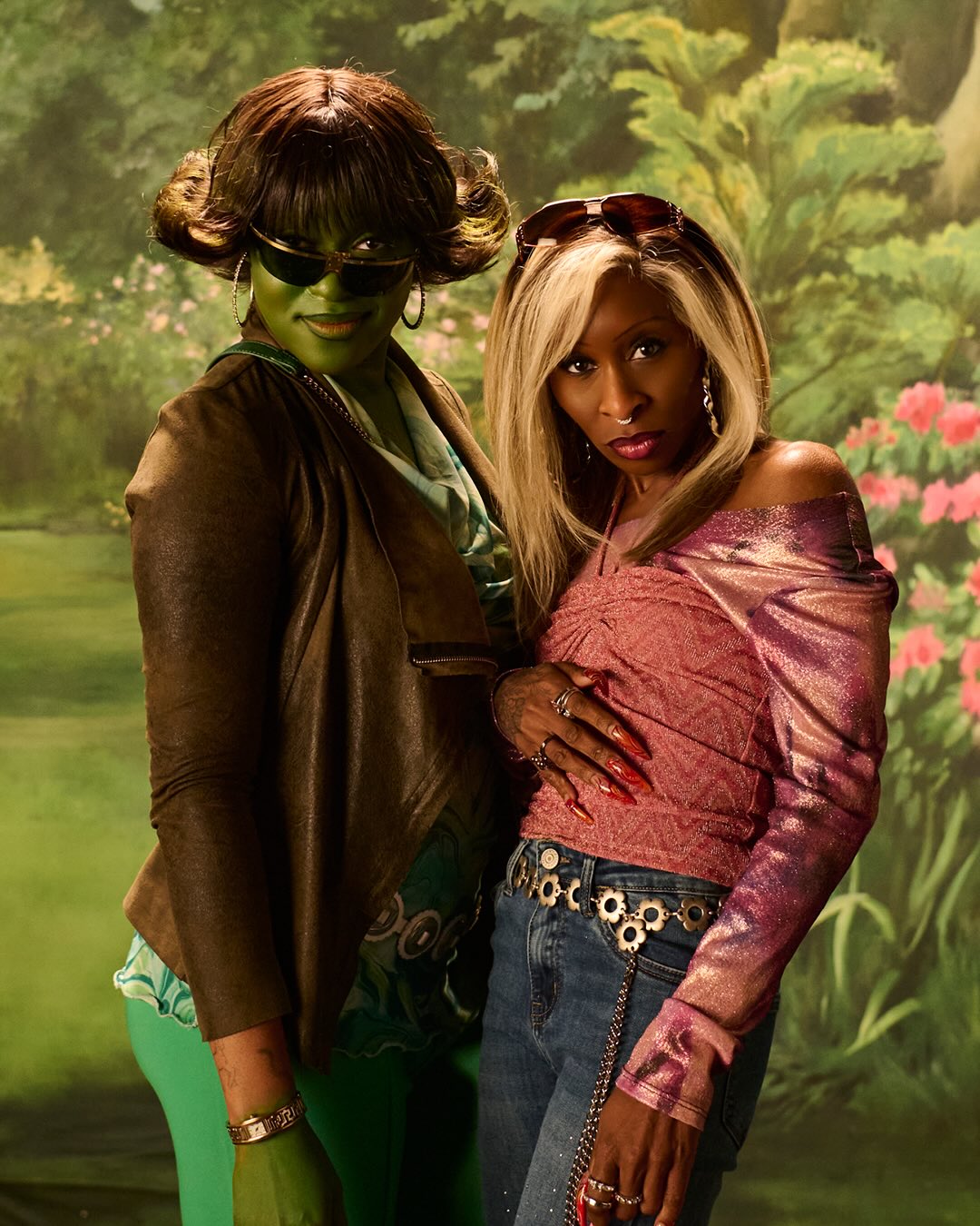 Cynthia Erivo wearing dark-rooted blonde side bangs with Ayoade Bamgboye in sunglasses and green body paint in front of a retro painted backdrop.