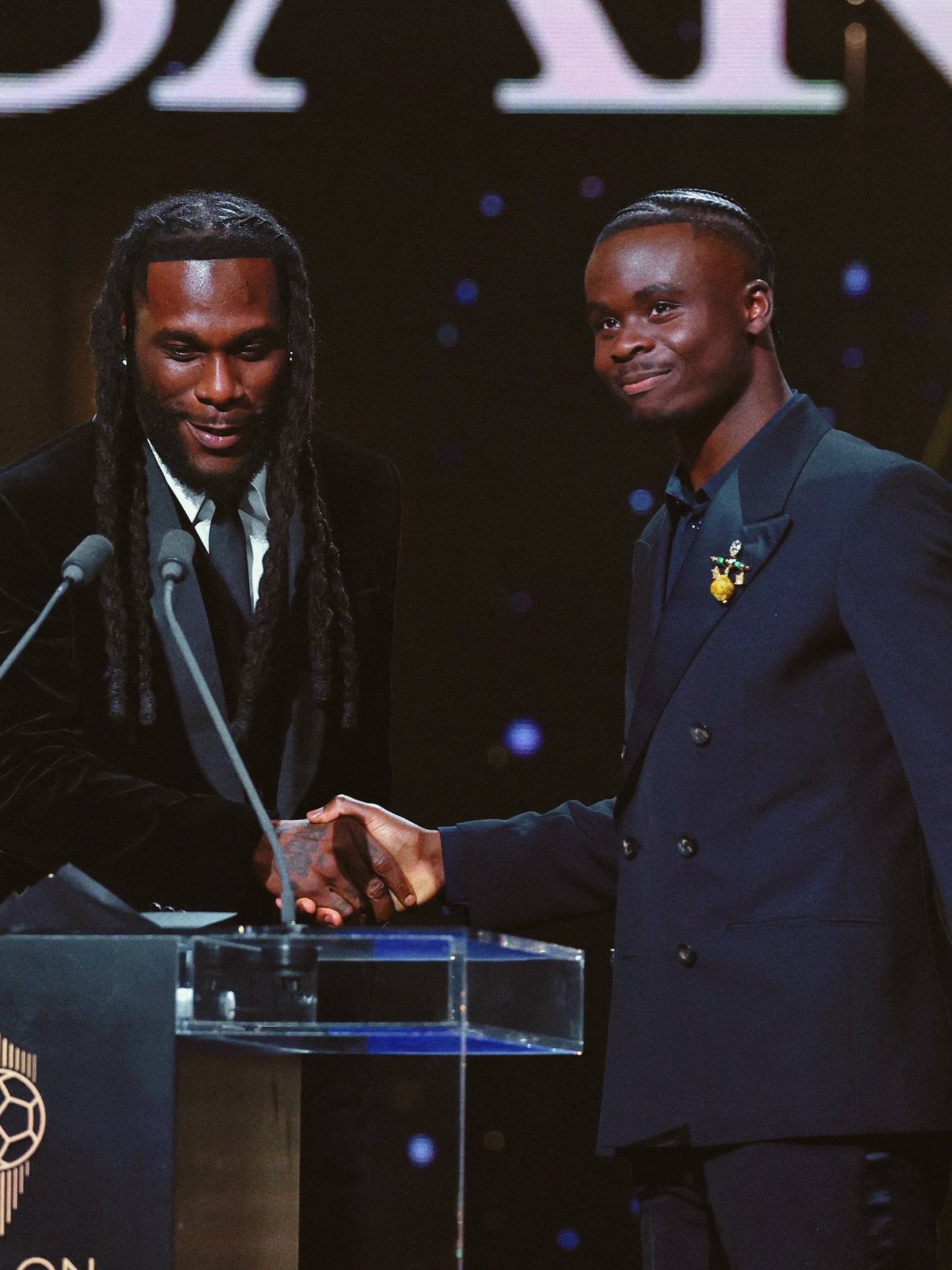 Burna Boy shaking hands with Bukayo Saka on stage at the 2026 Ballon d’Or ceremony.