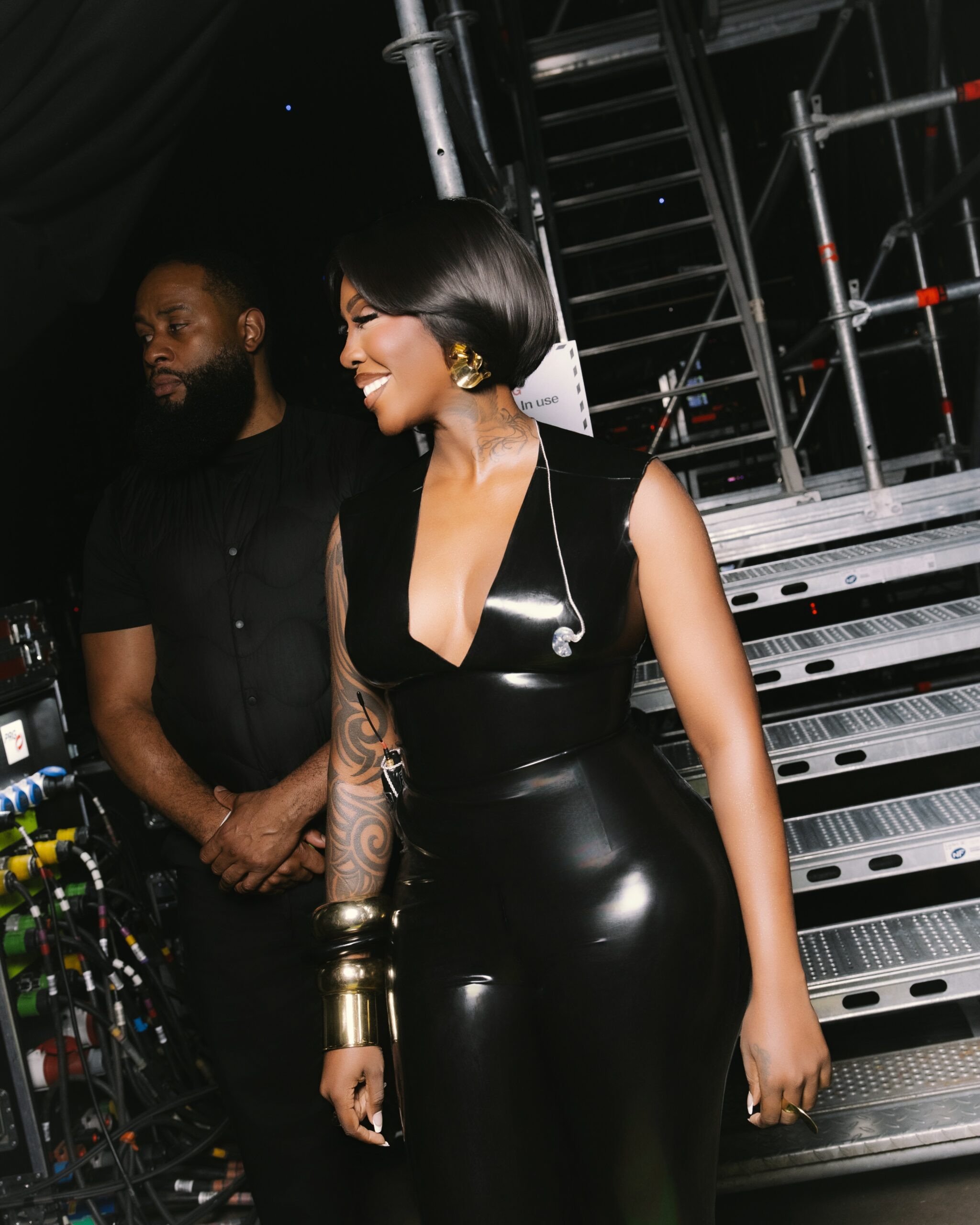 Tiwa Savage smiling backstage in a V-neck black latex catsuit with in-ear monitors.