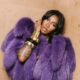 Tiwa Savage wrapped in a purple fur coat showing stacked gold bangles and glossy makeup.