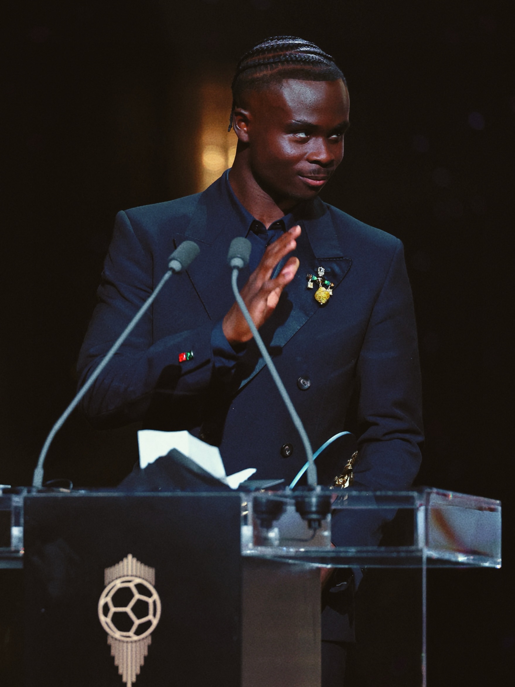 Bukayo Saka dressed in a black-tie suit clapping at the microphone stand after receiving the Socrates Award at the 70th Ballon d'Or ceremony.