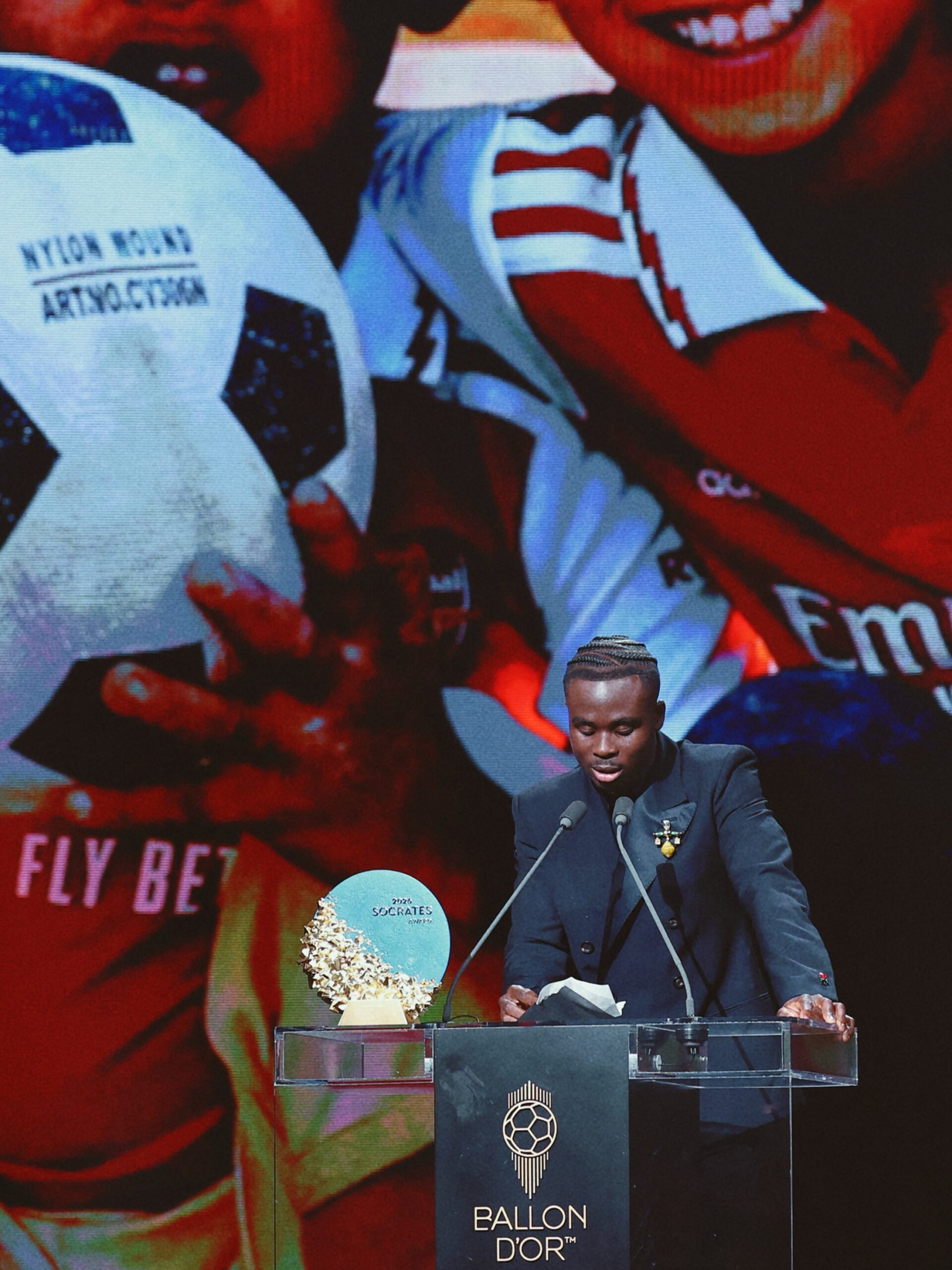 Bukayo Saka reading his speech at the podium with a background screen showing his charity work in Nigeria.