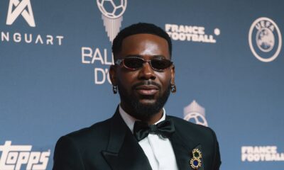 Adekunle Gold on the 2026 Ballon d'Or red carpet wearing a black double-breasted tuxedo, lapel brooch, and oval sunglasses.