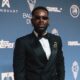 Adekunle Gold on the 2026 Ballon d'Or red carpet wearing a black double-breasted tuxedo, lapel brooch, and oval sunglasses.