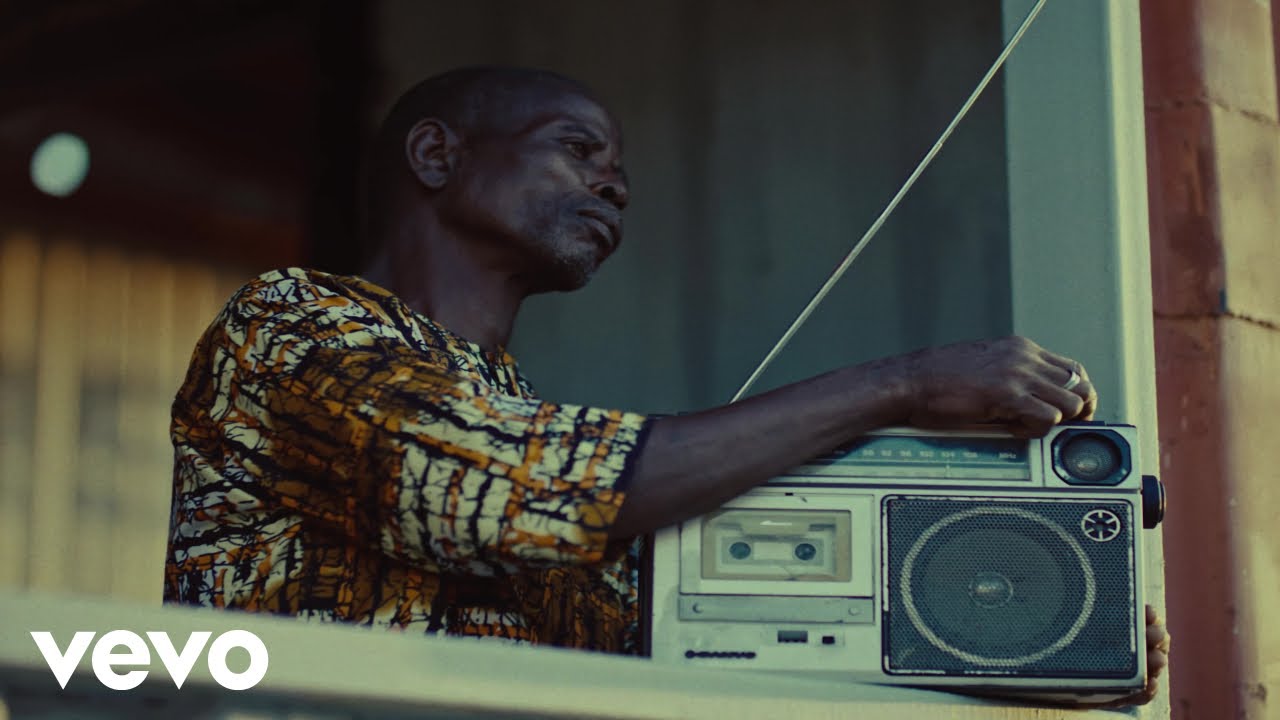 An elder wearing a patterned African print shirt tuning a silver boombox radio in Kizz Daniel Owo Oluwa music video